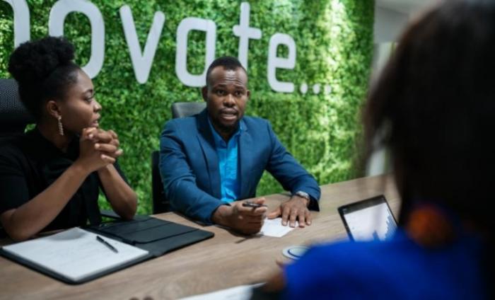 Business meeting between three people, with a green wall in the background that reads "Innovate" in white writing. 